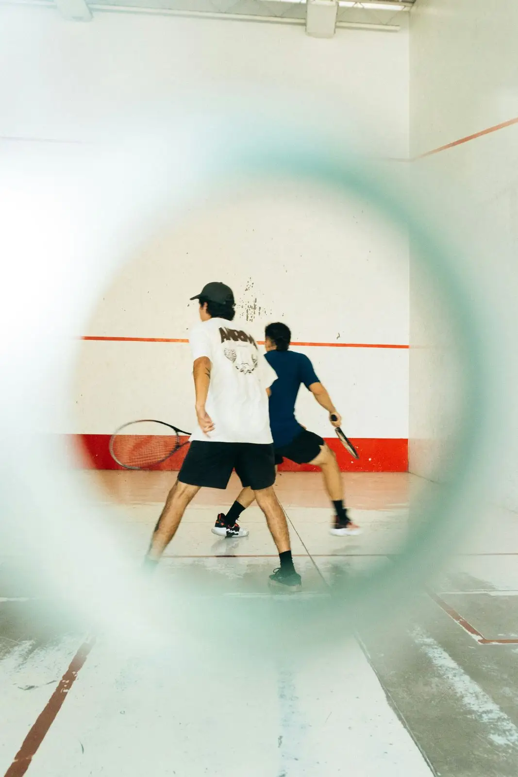 Two players mid-rally on a squash court, seen through a blurred ring